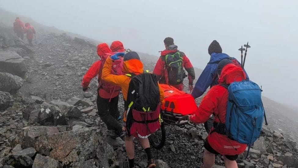 Members of Lochaber Mountain Rescue Team wheel a stretcher over rocky ground during an incident. The volunteers are wearing bright outdoor jackets.