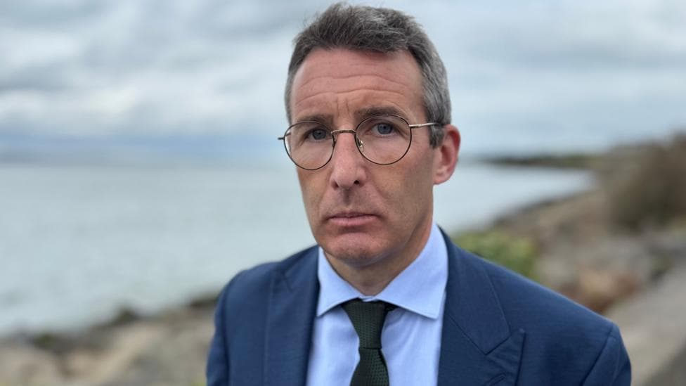 Andrew Muir, with short grey hair, wearing glasses, a blue shirt, black tie and navy suit.