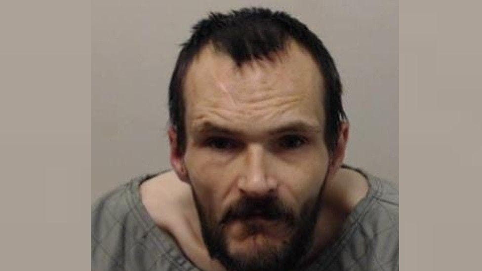 a police mugshot of a man with short scruffy brown hair and bear, wearing a grey wide-necked top
