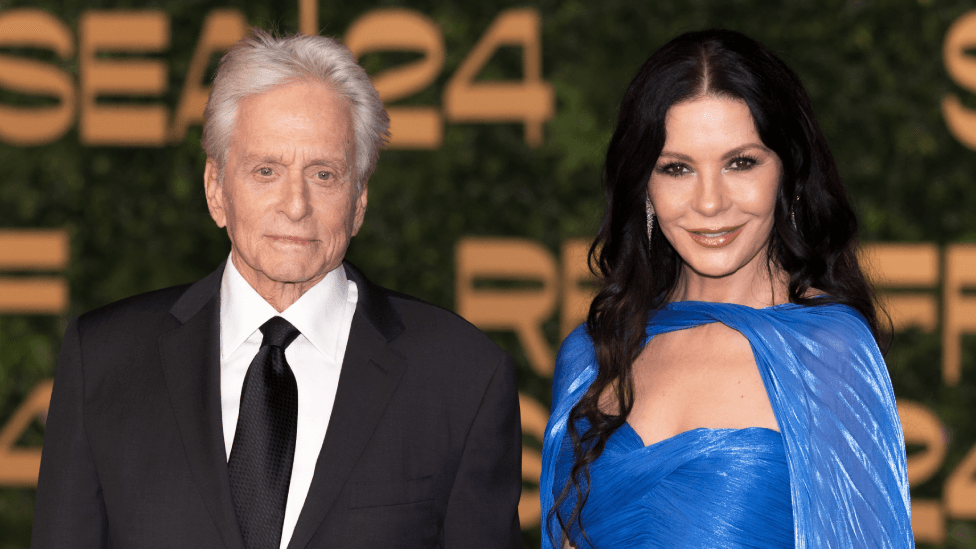 A man with white hair, wearing a tux and black tie stands next to a woman with long dark hair, wearing a strapless light blue ballgown, with matching chiffon cape.