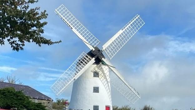 Picture of the windmill after it had been painted back to white. In front of the windmill is a red sign which reads 'Mr Holt's Mônuts'.