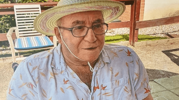 An old man sits in a chair and smiles. He is wearing glasses and has a straw hat on which is tied under his chin. He is wearing an open-necked shirt with a red and yellow pattern on it. He is outside and the sun is shining behind him.