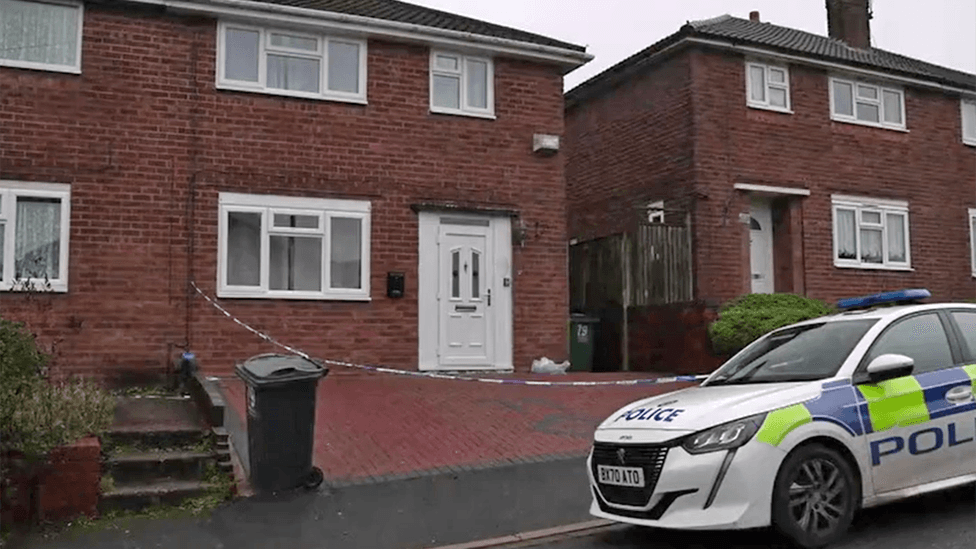 A police car is parked on the road outside a house, where police crime tape can be seen cordoning off the brick-paved driveway. The house has a white door and a white-framed front lower window.