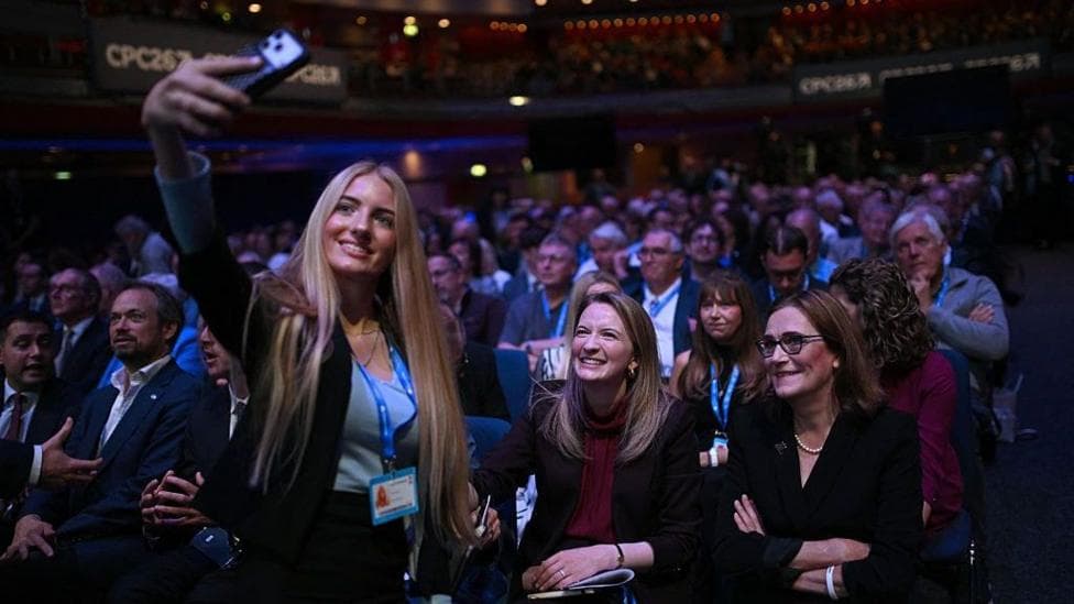 A young woman with long blonde hair takes a selfie with shadow housing secretary Katie Lam, who is sitting in the conference hall with Tory members