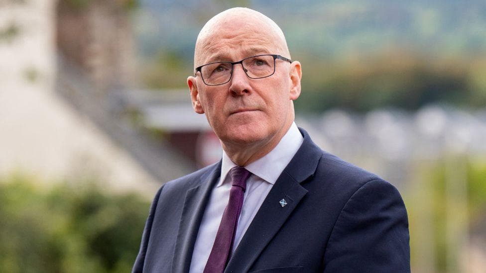 John Swinney, who is bald with glasses, gazing upwards. He is wearing a business suit