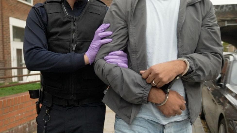A police officer wearing purple plastic gloves holds the arm of a man who is wearing handcuffs. they are walking along a pavement. The man is wearing a grey jacket, white t-shirt, light blue jeans and a ring on his left hand ring finger.