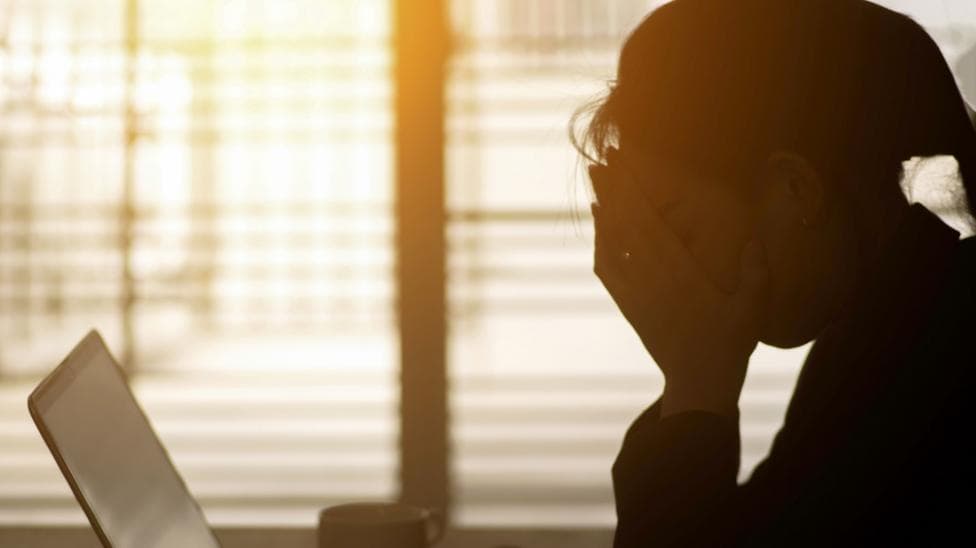 Stock image of woman with head in hands in front of computer. She is almost a silhouette. There is a cup and glasses sitting on the desk. There are blinds letting light in