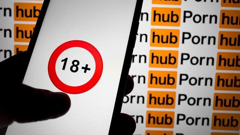 A hand holding a mobile phone with an 18+ logo with a red circle. The background is the Pornhub logo.
