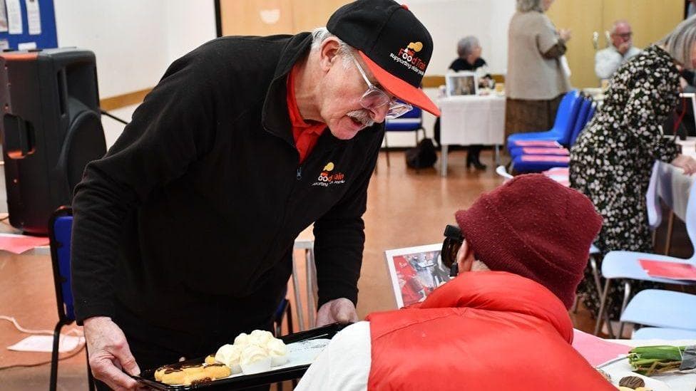 An old man with a moustache and glasses holding food on a tray