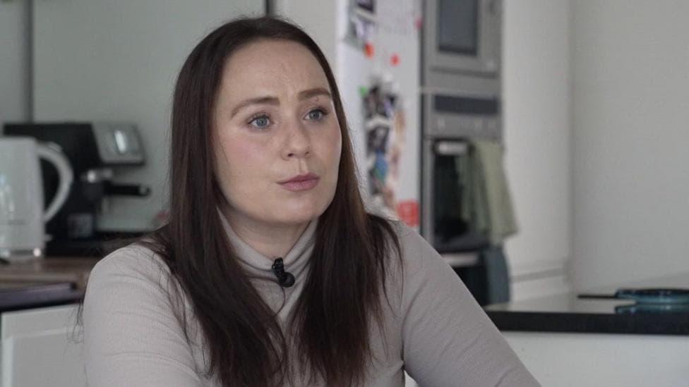 Rosanna Brown is sitting in a kitchen with the fridge and countertop out of focus behind her. She has long brown hair over her shoulders and is wearing a beige polo neck top.