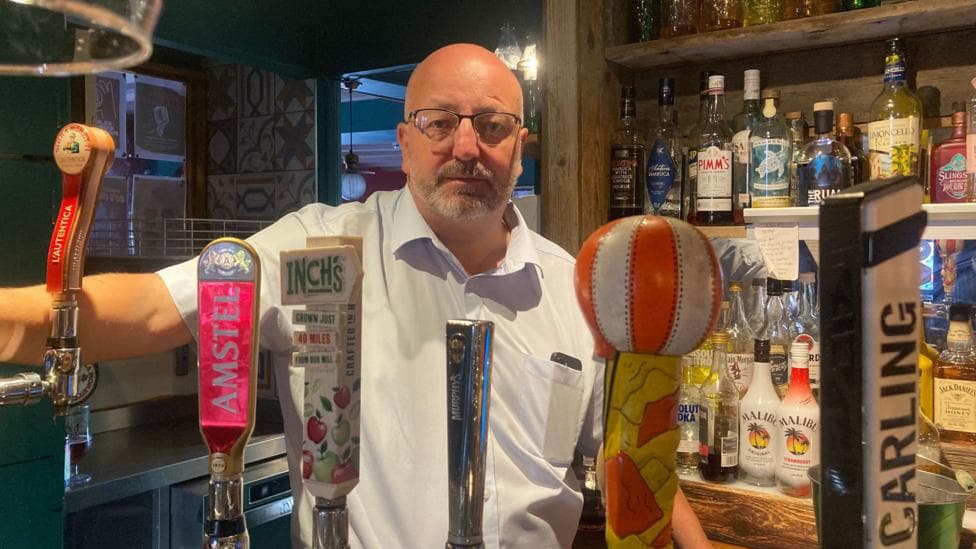 A man is standing behind a bar in a pub. He is wearing a short-sleeved light blue shirt and has one arm outstretched leaning on a bar pump in front of him.