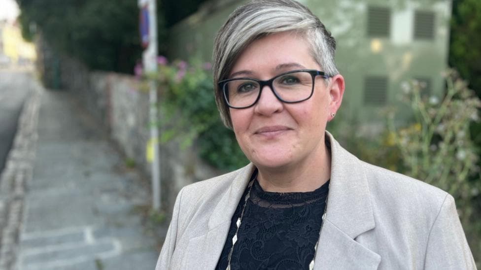 The image shows Deputy Haley Camp. She has short grey hair, longer on one side and swept over the top. She has black rimmed glasses and is wearing a grey blazer, with a black patterned blouse and a necklace with diamond-shaped beads on it.