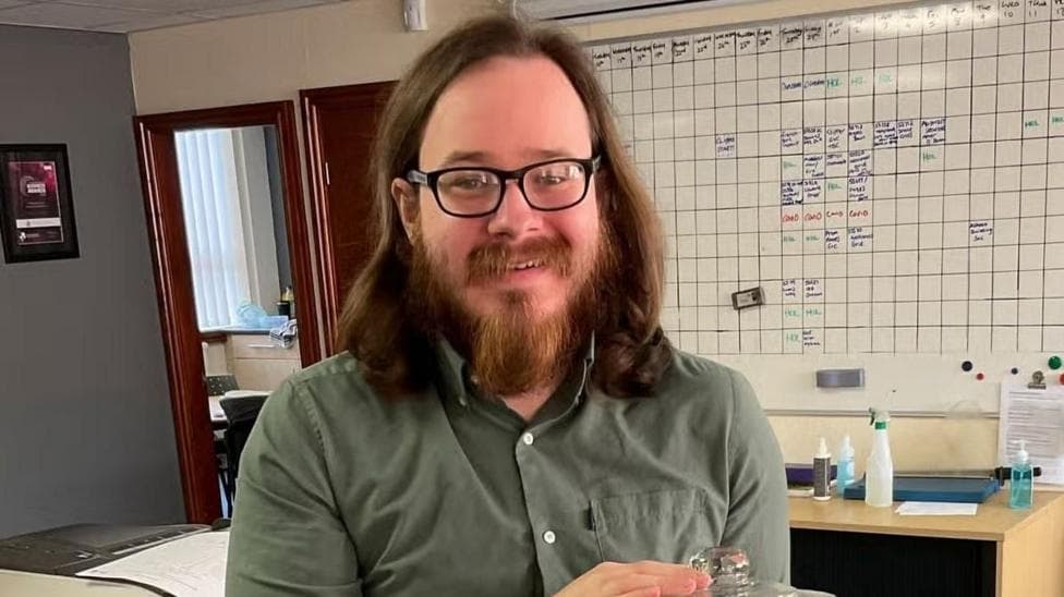 Joshua Kerry stands in a room with a whiteboard behind him. He wears a green shirt and glasses.