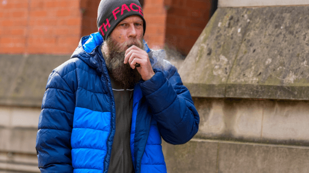A man in his 30s, with a large beard, grey woollen hat and blue bubble coat, stands outside a stone building holding a black vape to his mouth.