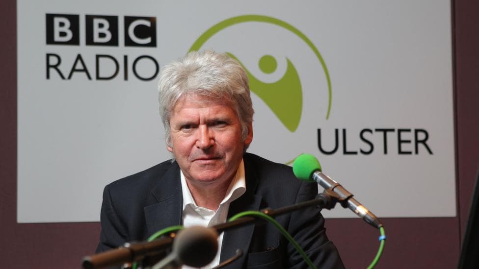 Man with grey hair, sitting in a radio ulster studio. There is a green coloured mic in front of him with a white radio ulster sign behind him. 'BBC radio' is written in black in the top right corner of the sign, with 'Ulster' written