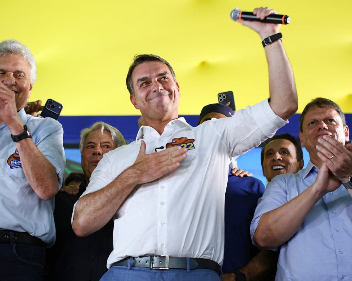 Flavio Bolsonaro raises a microphone, hand on chest, flanked by applauding men at a rally
