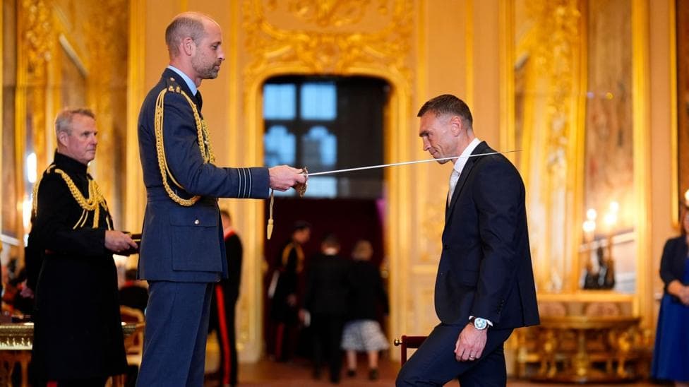 The Prince of Wales knights Sir Kevin Sinfield with a sword at an investiture in a grand, gold room at Windsor Castle.