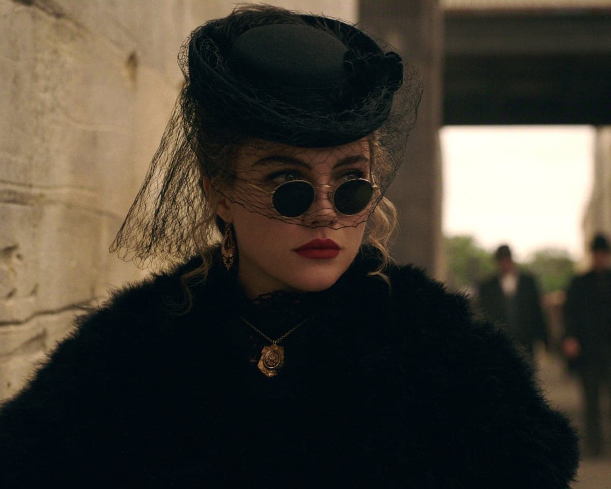 The actress seen outdoors by a wall looking kickass, dressed all in black, with round-rimmed black sunglasses