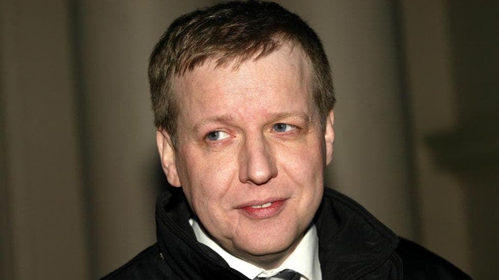 Paul Ferris, a pale man with a scarred face, is wearing a dark coat, white short and dark tie. He is smiling and looks off camera against a curtain backdrop.