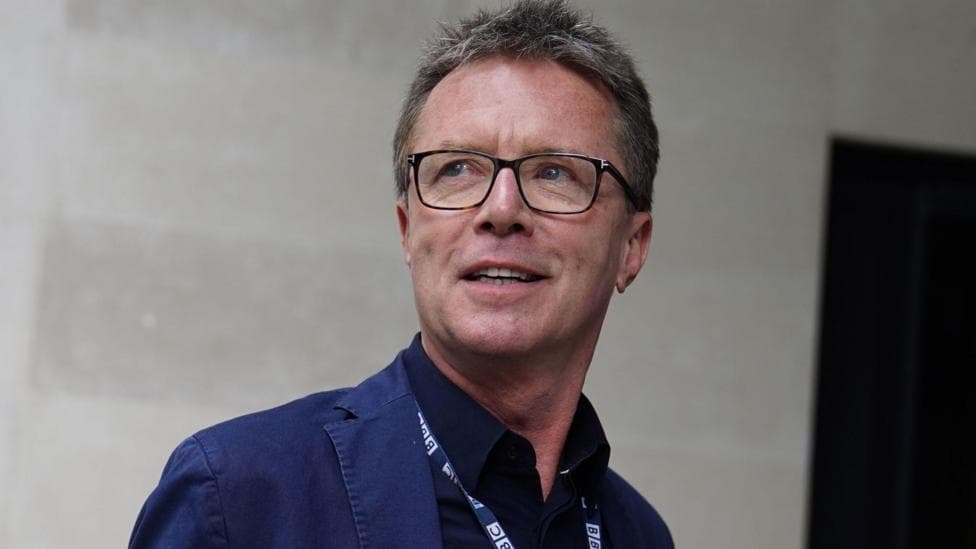 A man with grey spiky hair, black rectangular glasses, a blue blazer and a BBC lanyard