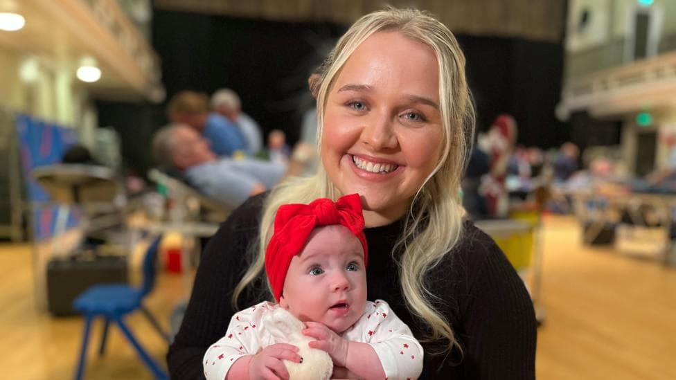 Caitlyn Stewart with blonde hair and wearing a black top smiles at the camera holding her daughter Millie, who is wearing a white and red baby grow and red bow headband.