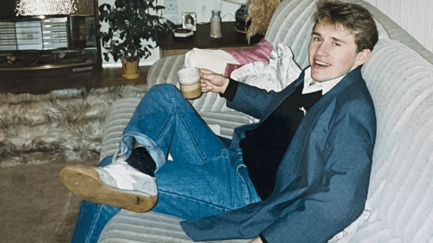 Anthony, a young man sits on a sofa. He is wearing a navy jacket and jeans and is holding a mug as he smiles at the camera. He has short brown hair and is clean shaven.