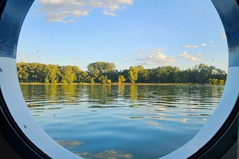 A photo taken out of a porthole on a river cruise vessel. Visible is the Dabune river with some trees in the background, against a blue sky.