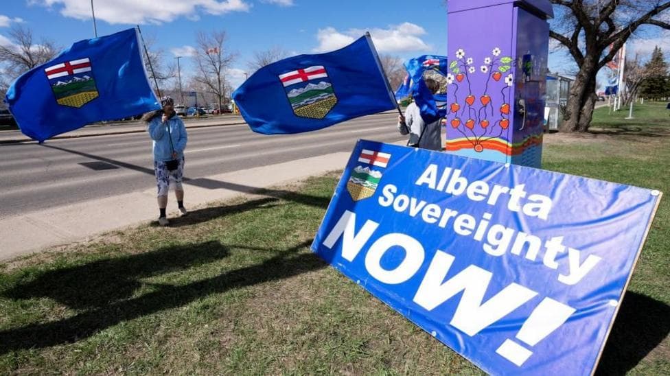 A man stands on a sidewalk and waves a blue Alberta flag. Propped up on the grass in front of him is a large sign in the same blue, with the provincial crest, that reads "Alberta Sovereignty Now!"