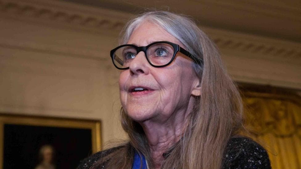 A woman with grey hair and black glasses looks upwards as she is being photographed.