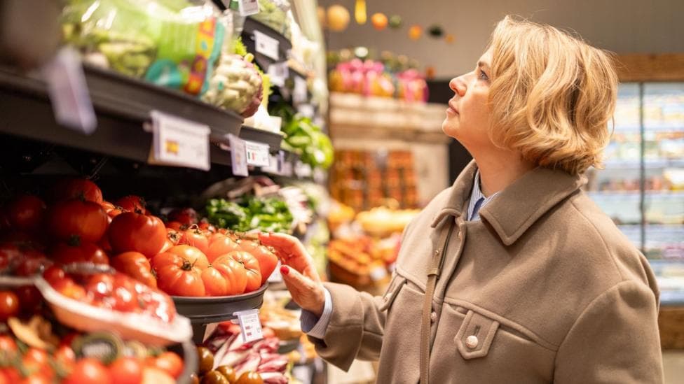 A woman with short blonde hair and wearing a beige coloured jacket is standing in a supermarket and looking at shelves that contain fruit and vegetables.