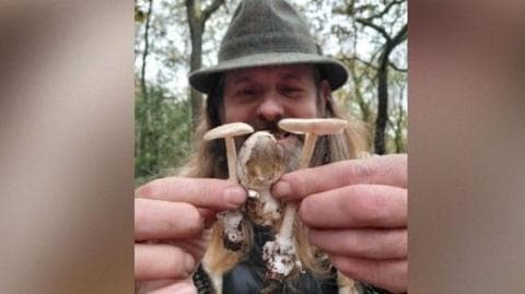 Colin Unsworth, wearing a dark outdoor hat, holds three olive amanita mushrooms, Amanita submembranacea, close to the camera with both hands. Two of the mushrooms have their pale gills visible beneath flat caps, while the smaller central mushroom has a rounded brownish cap. Woodland trees can be seen behind him.