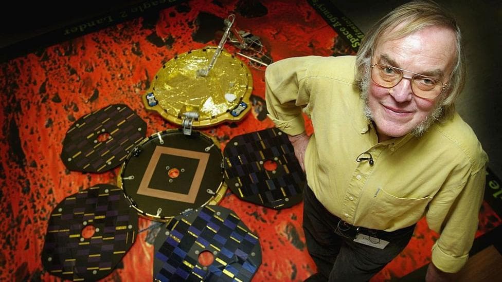 An older man with thick grey sideburns is stood next to a model of a Mars lander