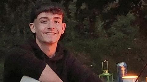 Daniel Colrain has short, dark hair. He is wearing a black, long-sleeved jumper and is sitting at an outdoor table at night time. There is a lamp and a can on the table