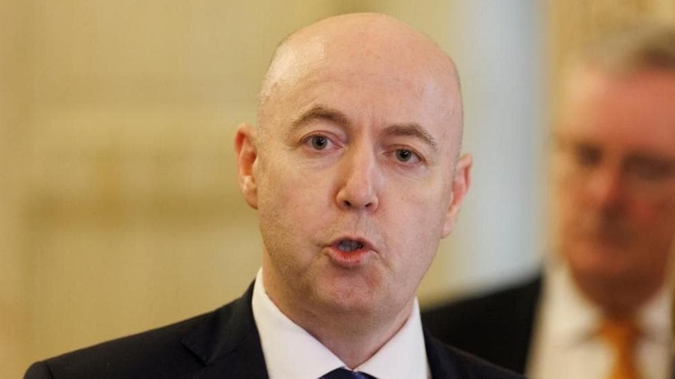 A headshot of Jon Burrows speaking. He's in Stormont's Great Hall and is wearing a black suit.