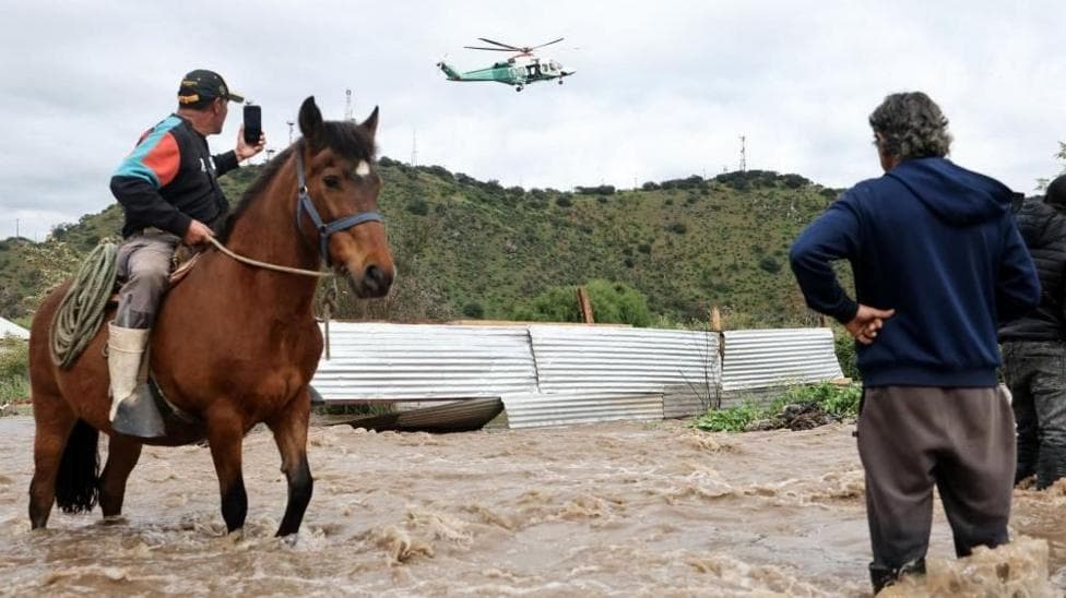 A man on a horse takes a video of a helicopter hovering overhead. He is wearing wellington boots and his horse is standing in water. Another man stands nearby, with his back to the camera. The water reaches his knees. The photo was taken in Talagante, in the Metropolitan Region of Chile, on 7 October 2026.