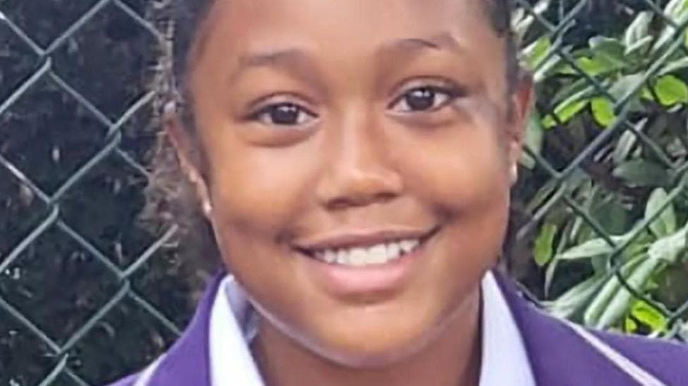 Mia St Hilaire, a 12-year-old in school uniform of a purple blazer and white shirt. She is standing in front of a chain-link fence and is smiling.