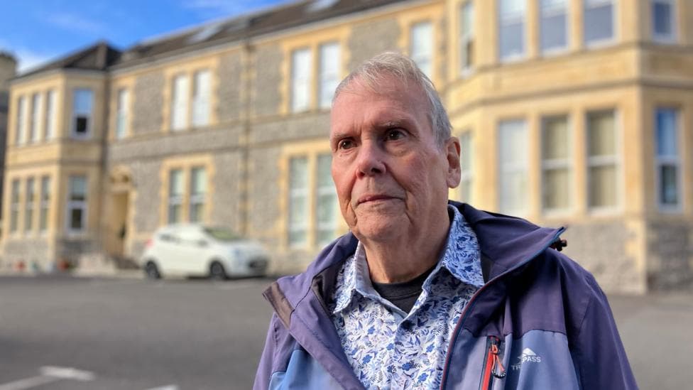 Alan, stands in the foreground of the photo. He has white hair. He wears a floral blue and white shirt with a rain coat over it. He stands in front of a large building which is in the background.