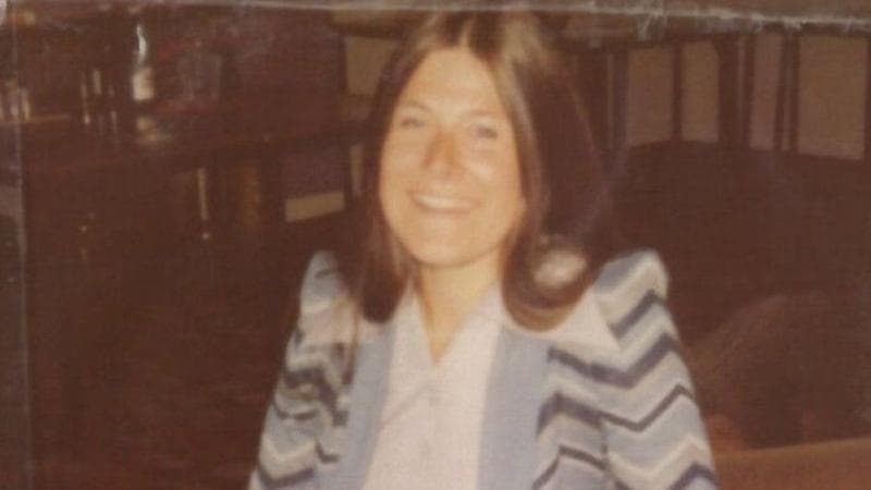 Brenda Page is pictured in the 1970s. She has shoulder length brown hair and is smiling as she sits in a room looking into the camera.