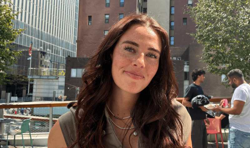 Willa, a young woman with long brown hair standing outside in front of buildings, smiling at the camera