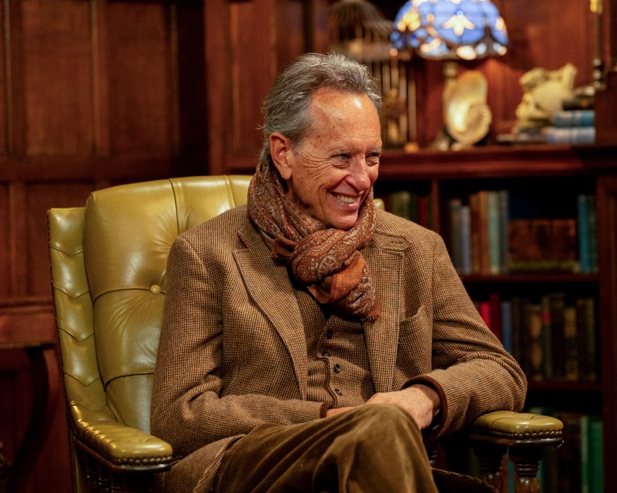 Richard E Grant smiles, seated in a leather armchair in a wood-panelled library