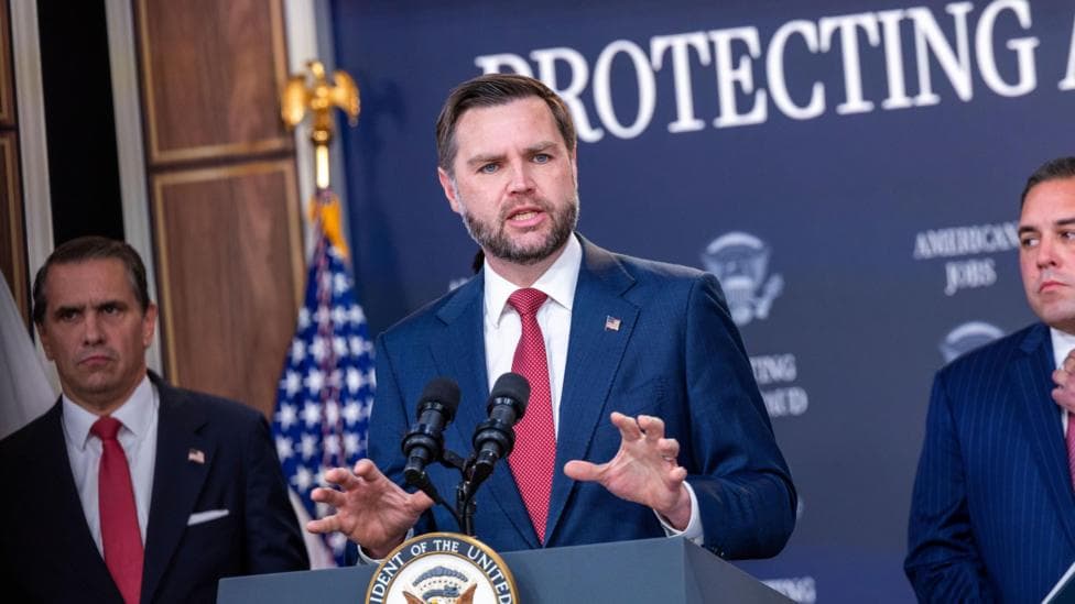 Vice President JD Vance standing at a podium and gesturing, wearing a blue suit, white shirt, and red necktie.