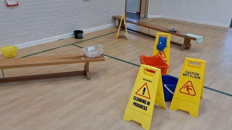 A number of wet floor signs are visible in an assembly hall with a number of buckets on benches,