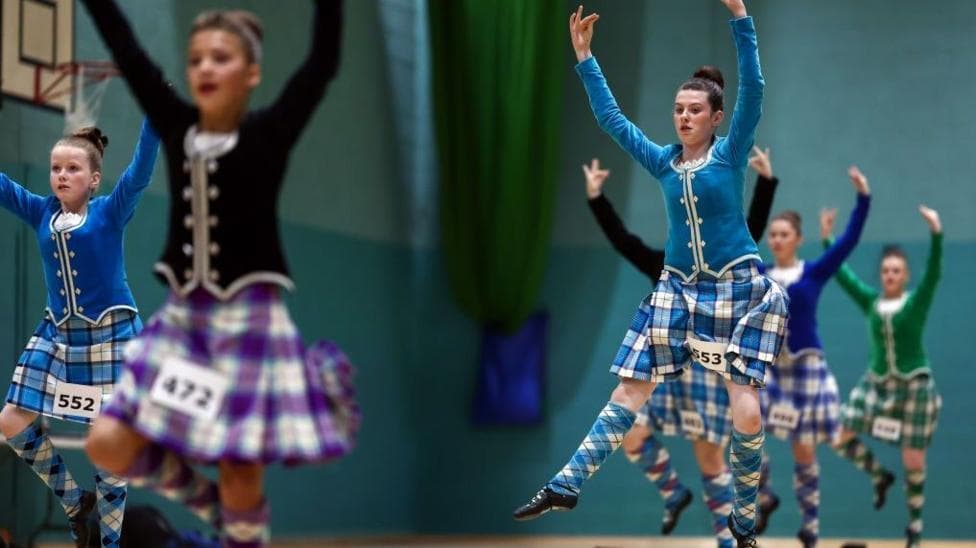 Six young Highland dancers, dressed in blue, green or purple traditional dress, leap from the floor of a sports hall while competing in the Mòd.