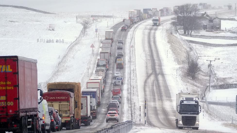 A road covered with snow and ice with snowy fields on either side, with a queue of cars and lorries in one direction and two lorries moving in the other.