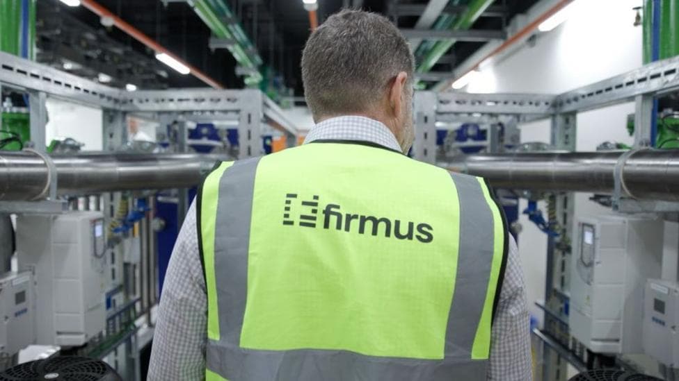 A man in a Firmus-branded safety jacket inspects racks of computer systems in a data centre.