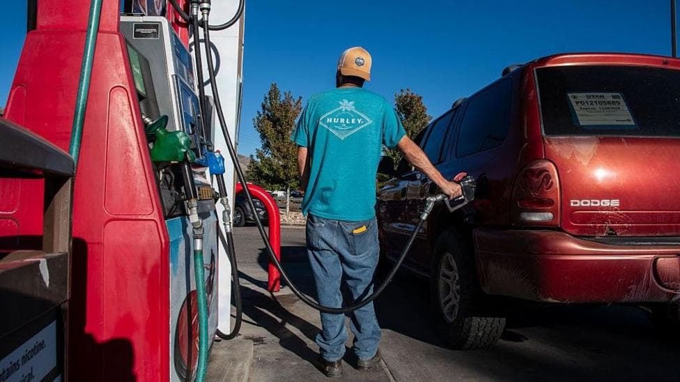 A customer wearing a backwards cap and a blue tshirt refuels a red vehicle at a gas station in Salt Lake City, Utah, United States on a clear, sunny day.