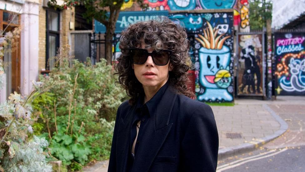 Meredith Whittaker wearing dark sunglasses and a black jacket, standing outside a cafe, in front of colourful street art. She has dark, curly, shoulder-length hair with a fringe.