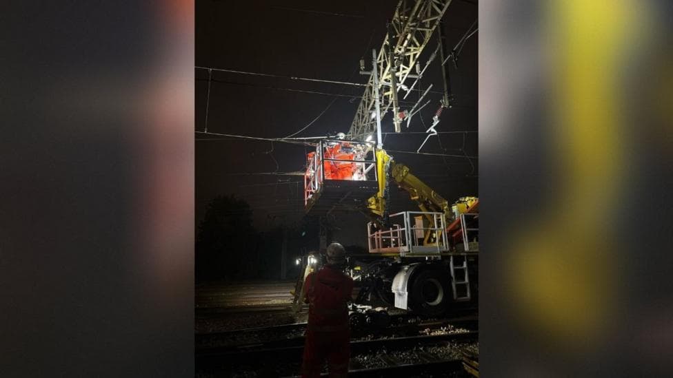 Railway engineers wearing orange high-visibility protective clothing and white safety helmets work from a raised metal platform beside overhead electrical wires at night.