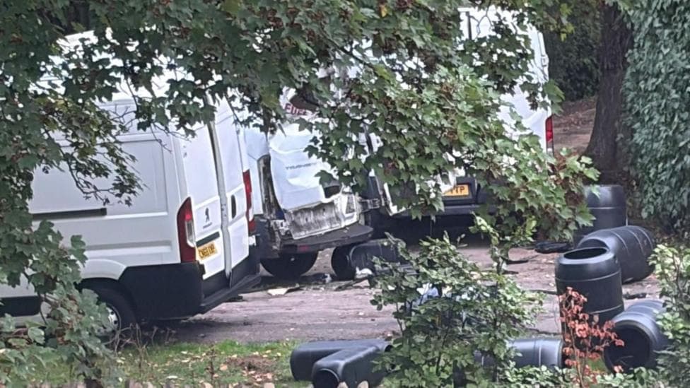 Three white vans with a load of black barrels out the back. One of the vans has its doors blown off.