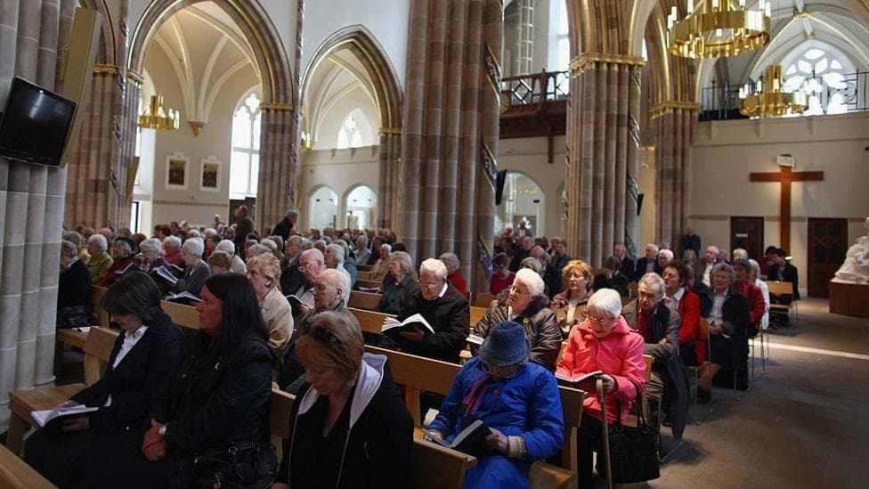 The inside of a Glasgow church, with parishioners sitting on pews and singing from hymn book. A large cross is visible on the back wall, with large pillars sitting between the pews.